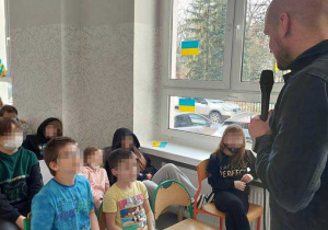 Dzieci siedzą na krzesełkach zwrócone w stronę przemawiającego mężczyzny. Na twarzach dzieci rysuje się zaciekawienie. Mężczyzna trzyma w ręku mikrofon i przemawia stojąc przed dziećmi